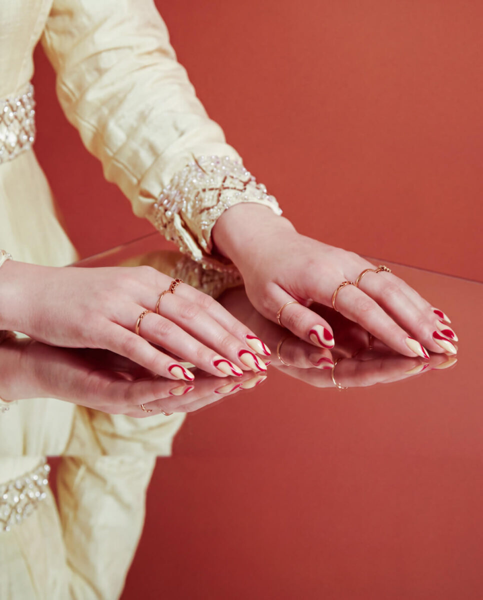 Almond nails with cream and deep red swirl nail art, hands resting on a mirrored surface against a terracotta background